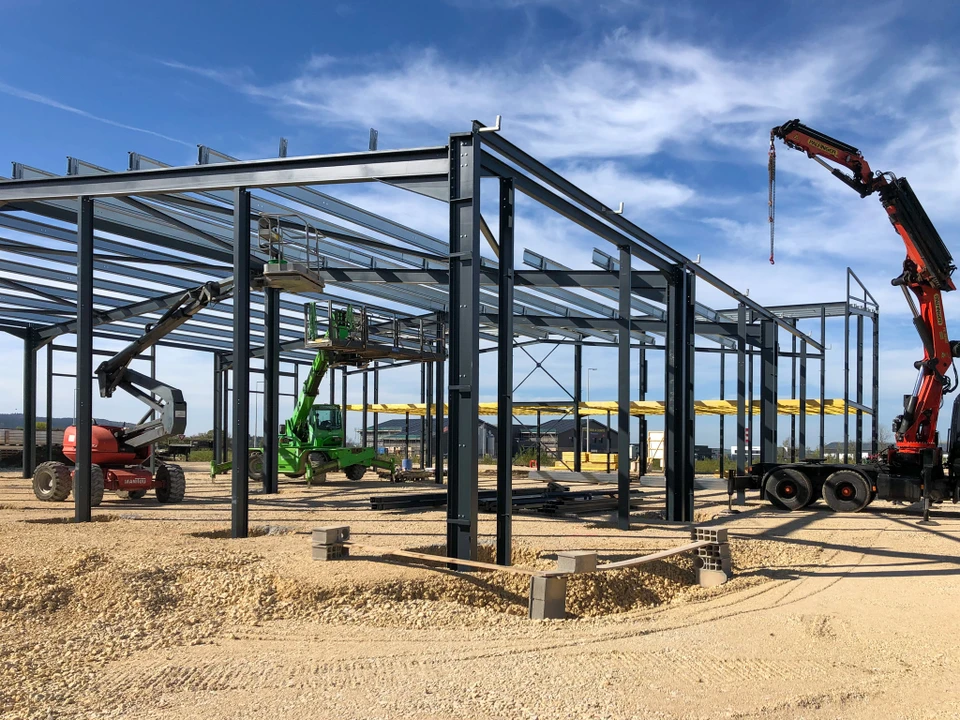 Un professionnel en tenue de travail (casque, gilet de sécurité) discutant avec un client sur un chantier de construction métallique, examinant ensemble des plans de projet. En arrière-plan, une structure métallique en cours de montage avec charpente et ossature en acier visible. L'image doit transmettre la confiance, le professionnalisme et la relation client personnalisée dans un contexte de construction métallique en Haute-Saône.